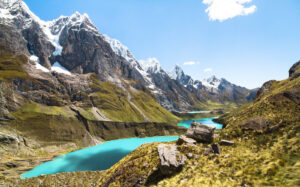 A scenic view of a mountain range with snow-capped peaks under a clear blue sky. In the foreground, there is grassy terrain and large rocks, perfect for a hiking tour. Turquoise lakes are nestled between the mountains, reflecting the vibrant color of the sky.