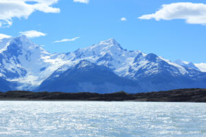 A breathtaking view of snow-capped mountains under a bright blue sky with scattered clouds. The mountains reflect in a clear, shimmering body of water in the foreground, with a dark rocky shoreline visible between the water and the mountains—an ideal setting for an unforgettable hiking tour.