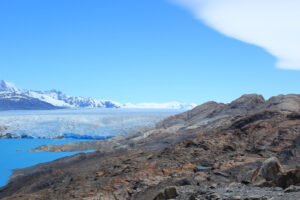 A striking landscape showing a vast glacier extending into the distance with snow-covered mountains in the background. The foreground features rocky terrain leading to turquoise water of a glacial lake under a clear blue sky, perfect for an unforgettable hiking tour.