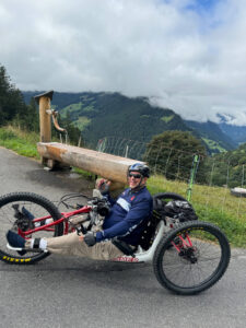 A person riding a recumbent tricycle on a mountain road, smiling and showing a thumbs-up. They are wearing sunglasses, a helmet, and outdoor gear. A wooden water spout and lush, cloudy mountains are visible in the background. .