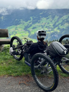 A person wearing a helmet is seated on a recumbent three-wheeled bicycle, looking out at a scenic view of green mountains and valley under a cloudy sky. The bicycle is parked near a wooden bench on a grassy area beside a paved trail.