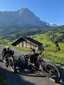 Two people on all-terrain handcycles smile while posing on a paved road with a backdrop of green hills, a rustic wooden barn, and towering mountains under a clear blue sky. One person raises a hand, celebrating the moment. Flowers and trees dot the landscape.