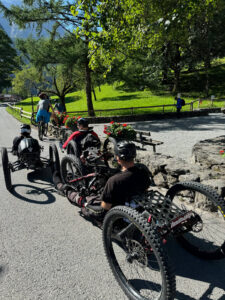 A group of people with helmets ride on adaptive mountain bikes down a paved path in a scenic outdoor area surrounded by trees and greenery. Some people are seen walking nearby, and there's a stone fence with flower boxes alongside the path.