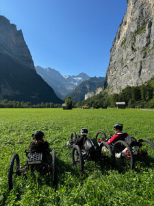 Three individuals are sitting in adaptive off-road vehicles in a lush green field, surrounded by towering mountains under a clear blue sky. They are wearing helmets and appear to be taking in the scenic views of the mountain landscape.