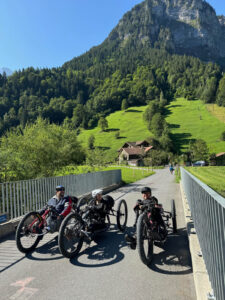 Three people on handcycles are positioned on a bridge with a lush, green mountainous landscape in the background. The sky is clear blue, and a quaint house is nestled among the trees and grassy hills.