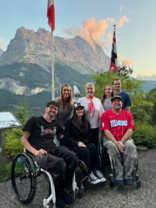 A group of seven people, including two individuals in wheelchairs, pose together outdoors with a scenic mountain and valley backdrop. They are smiling and surrounded by greenery, with a flagpole and informational sign nearby. The sky is clear with a few clouds.