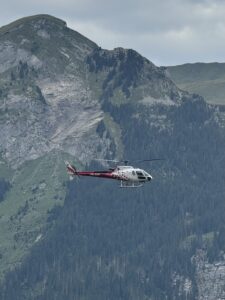 A helicopter with a red and white color scheme flies in front of a mountainous landscape. The scene features rugged, forested slopes and rocky cliffs under a cloudy sky.