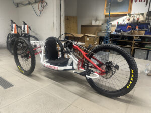 A sleek, white off-road handcycle with large, rugged tires is positioned in a workshop. The brand "Lasher" is displayed on its frame. Various tools, equipment, and bicycle components are visible in the background.