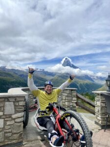 Person in a yellow jacket and helmet smiling and posing with thumbs up while sitting on a handcycle. They are on a terrace with a stone railing, with the Matterhorn mountain and cloudy sky in the background.