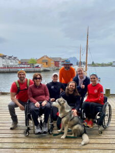 A group of seven smiling people, two in wheelchairs, and a dog sit and stand on a wooden dock by the water, with colorful houses and a sailboat in the background.
