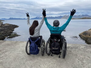 Two people in wheelchairs face a calm body of water, raising their arms in celebration. They wear winter clothing and backpacks, surrounded by rocky shores and distant mountains under a cloudy sky.