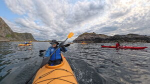 A group of kayakers paddles on calm water surrounded by rocky hills under a cloudy sky. The photo is taken from a yellow kayak with a person in blue gear and a hat smiling at the camera.
