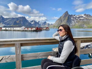 A woman wearing sunglasses and a black vest sits on a wooden railing, gazing at a scenic fjord with mountains, colorful houses on the water, and partly cloudy skies in the background.
