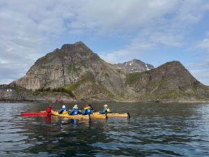 A group of people kayaking in yellow and red kayaks on calm water, with steep, rocky mountains and a partly cloudy sky in the background.