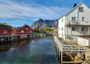 A scenic waterfront view with red and white wooden houses on stilts over clear water, rocky shoreline, and distant mountains under a blue sky with wispy clouds.