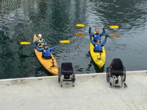 Two yellow kayaks with two paddlers each, all wearing life jackets, float on calm water near a dock. Two empty wheelchairs are parked on the dock in front of the kayakers, who are smiling and raising their paddles.
