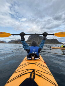 A person in a yellow kayak raises their paddle overhead while facing a scenic view of mountains and water under a cloudy sky. Another kayaker is visible in the distance on the right.