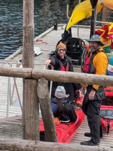Three people prepare for kayaking on a dock; one man in sunglasses and a beanie is holding a paddle and smiling up, another man in a hat and yellow jacket stands beside a seated person in a red kayak.