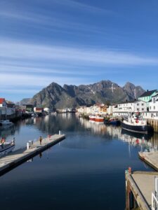 A calm harbor with boats docked along both sides, colorful houses lining the waterfront, and mountains with patches of snow in the background under a blue sky.
