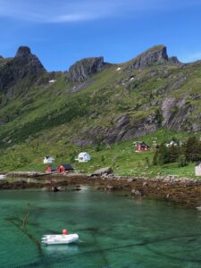 A small white boat floats on clear turquoise water near a rocky shore. Colorful houses sit on green hills beneath rugged, steep mountains under a bright blue sky.