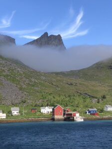 A red house sits by the water with green hills and scattered houses behind it. Fog partially covers the hillside, and a rugged mountain peak rises above under a bright blue sky with wispy clouds.