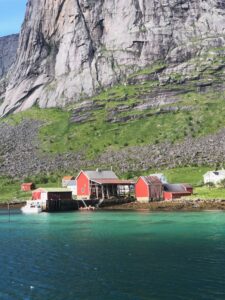 Red cabins sit by clear turquoise water at the base of a steep, rocky mountain, with patches of green grass and a bright, sunny sky.