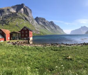 A bright red wooden house stands by a calm fjord, surrounded by green grass and rocky mountains under a clear blue sky. Other red buildings are nearby, and the landscape is peaceful and scenic.