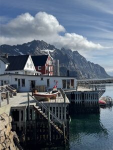 Wooden houses, including a red and a white one, stand on stilts over clear water with small boats docked nearby. Rocky mountains rise in the background under a partly cloudy sky.