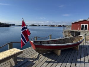 A wooden boat with a Norwegian flag sits on a dock beside a calm bay. A red building stands nearby, and small islands and houses are visible across the water under a blue sky.