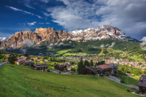 A picturesque alpine village with wooden houses nestled in a green valley, surrounded by lush trees and dramatic mountain peaks under a partly cloudy sky.
