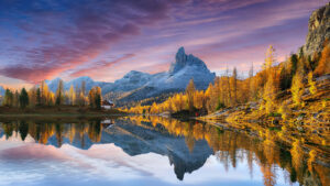 A serene mountain landscape at sunrise, with snow-capped peaks, golden autumn trees, and a calm lake reflecting the scenery. A small house and a person in red stand near the water’s edge.