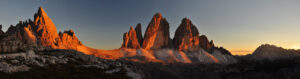 Panoramic view of rugged mountain peaks glowing orange in the light of a setting or rising sun, with rocky foreground and a clear sky in the background.