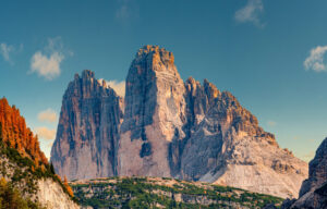 Massive, rugged mountain peaks rise against a clear blue sky, surrounded by green forests and autumn-colored trees on the left. Sunlight highlights the rocky texture and dramatic cliffs of the landscape.
