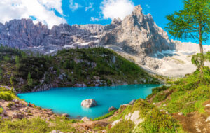 A vibrant turquoise alpine lake surrounded by rocky mountains and green slopes, with a tall tree on the right and patches of snow above the water, beneath a bright blue sky with some clouds.