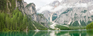 A serene mountain lake surrounded by dense green pine forests and tall rocky peaks partially covered by clouds, with a few small boats floating on the calm water.